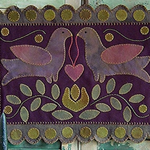 Peut inclure: Une tenture murale en feutre faite à la main représentant deux oiseaux se faisant face, reliés par un cœur rose. Les oiseaux sont violets avec des détails roses, sur un fond violet foncé. La bordure est grise avec des cercles verts.
