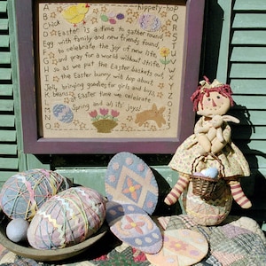 Op de afbeelding: Een ingelijst borduurwerk met een alfabet en een gedicht over Pasen. Het gedicht luidt: "A Chick hippety-hop Easter is a time to gather round with family and new friends found, Egg to celebrate the joy of new life, and pray for a world without strife, so as we put the Easter baskets out, the Easter bunny will hop about, Jelly bringing goodies for girls and boys, beans at Easter time we celebrate, Spring and all its joys!" Het borduurwerk is op een groene houten achtergrond met een lappenpop die een mand met paaseieren vasthoudt.