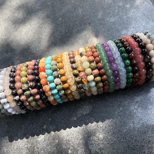 Op de afbeelding: Een verzameling kralenarmbanden in verschillende kleuren, waaronder wit, zwart, turquoise, oranje, geel, groen, paars en roze. De armbanden worden gepresenteerd op een grijze cilindrische houder met gouden accenten, met een verscheidenheid aan natuurstenen kralen.