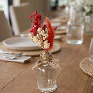 Peut inclure: Un petit vase en verre transparent, avec des rainures verticales, noué avec de la ficelle et un nœud, contient un bouquet de fleurs séchées. L'arrangement comprend des éléments rouges, crème et orange, posé sur une table en bois. Une décoration de table.