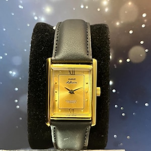 Könnte beinhalten: Eine rechteckige Armbanduhr in Goldoptik mit einem schwarzen Lederarmband. Das Zifferblatt der Uhr ist goldfarben mit schwarzen Zeigern und römischen Ziffern. Die Uhr ist mit "Futle Ashvini" und "Parashock 17 Jewels" beschriftet.