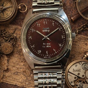 Könnte beinhalten: Eine Vintage-Armbanduhr mit braunem Zifferblatt und silberfarbenem Metallarmband. Das Zifferblatt der Uhr zeigt arabische Ziffern und Text in einer Fremdsprache. Weitere antike Gegenstände, darunter ein Kompass und eine Lupe, befinden sich im Hintergrund.