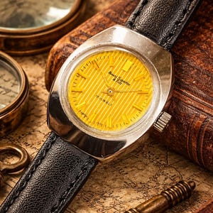 Könnte beinhalten: Eine Vintage-Armbanduhr mit silbernem Gehäuse und einem hellgelben, gestreiften Zifferblatt. Die Uhr hat ein schwarzes Lederarmband und der Text "Henry Sandoz & Fils" ist auf dem Zifferblatt zu sehen. Die Uhr befindet sich vor dem Hintergrund antiker Karten und ledergebundener Bücher.