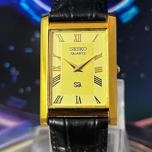 May include: A rectangular Seiko Quartz wristwatch with a gold-tone case and a yellow dial featuring black Roman numeral hour markers. The watch has a black leather strap with a textured finish and gold-tone hands for the hour, minute, and second.