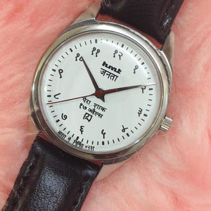 May include: A silver-toned wristwatch with a white face and black leather band. The watch face features black numerals, black hands, and text in a foreign language. The watch is set against a pink, fuzzy background.