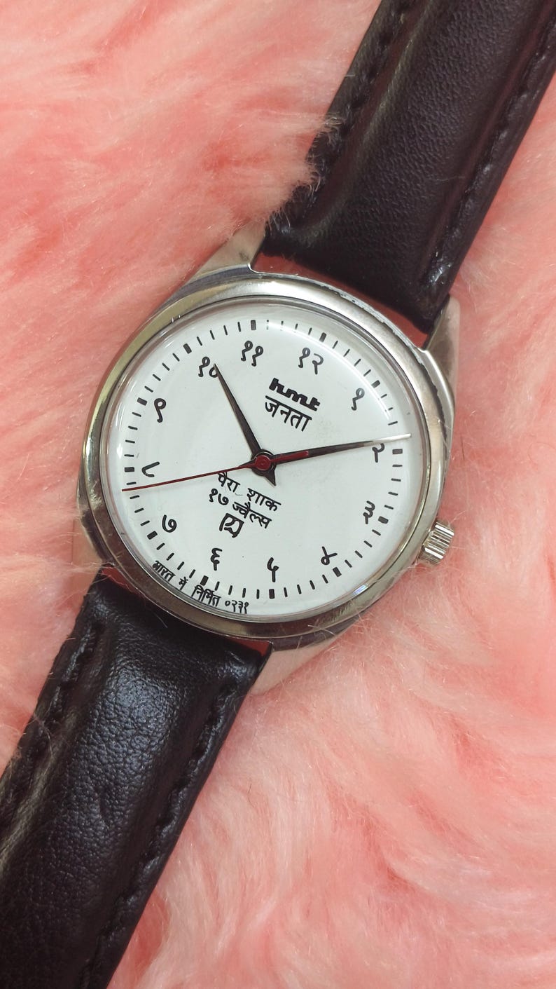 May include: A silver-toned wristwatch with a white face and black leather band. The watch face features black numerals, black hands, and text in a foreign language. The watch is set against a pink, fuzzy background.
