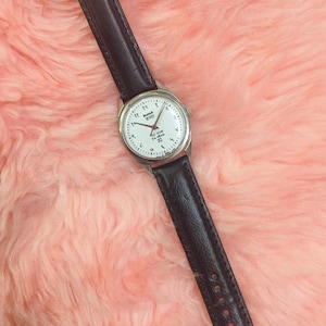 May include: A wristwatch with a white face, silver casing, and black leather band. The watch face features black numerals and hands. The watch is set against a pink, fluffy background.