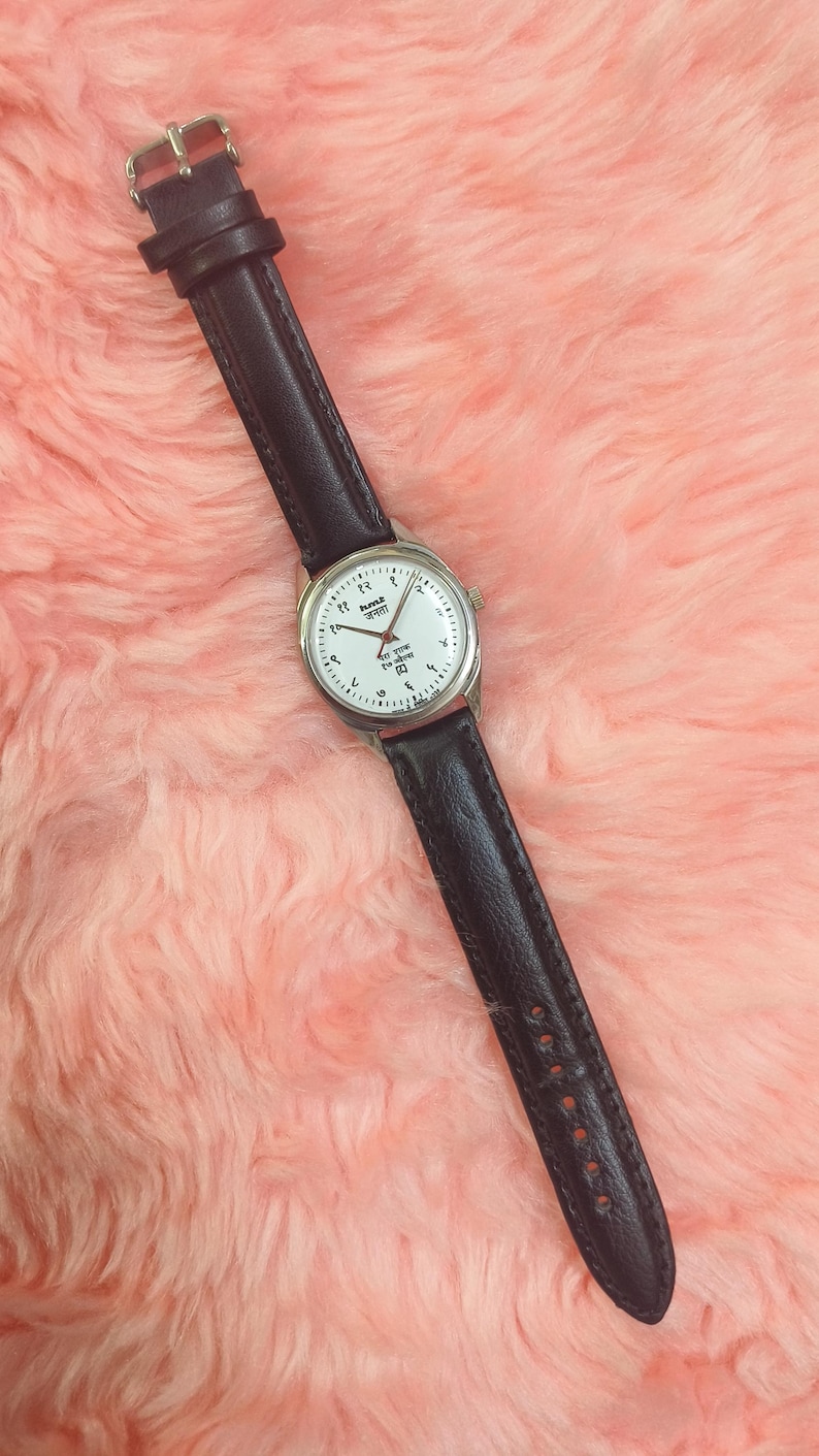 May include: A wristwatch with a white face, silver casing, and black leather band. The watch face features black numerals and hands. The watch is set against a pink, fluffy background.