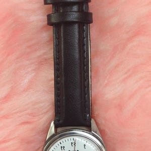 May include: A wristwatch with a silver-colored case and a black leather strap. The watch face is white with black numerals and text. The watch has a buckle closure.