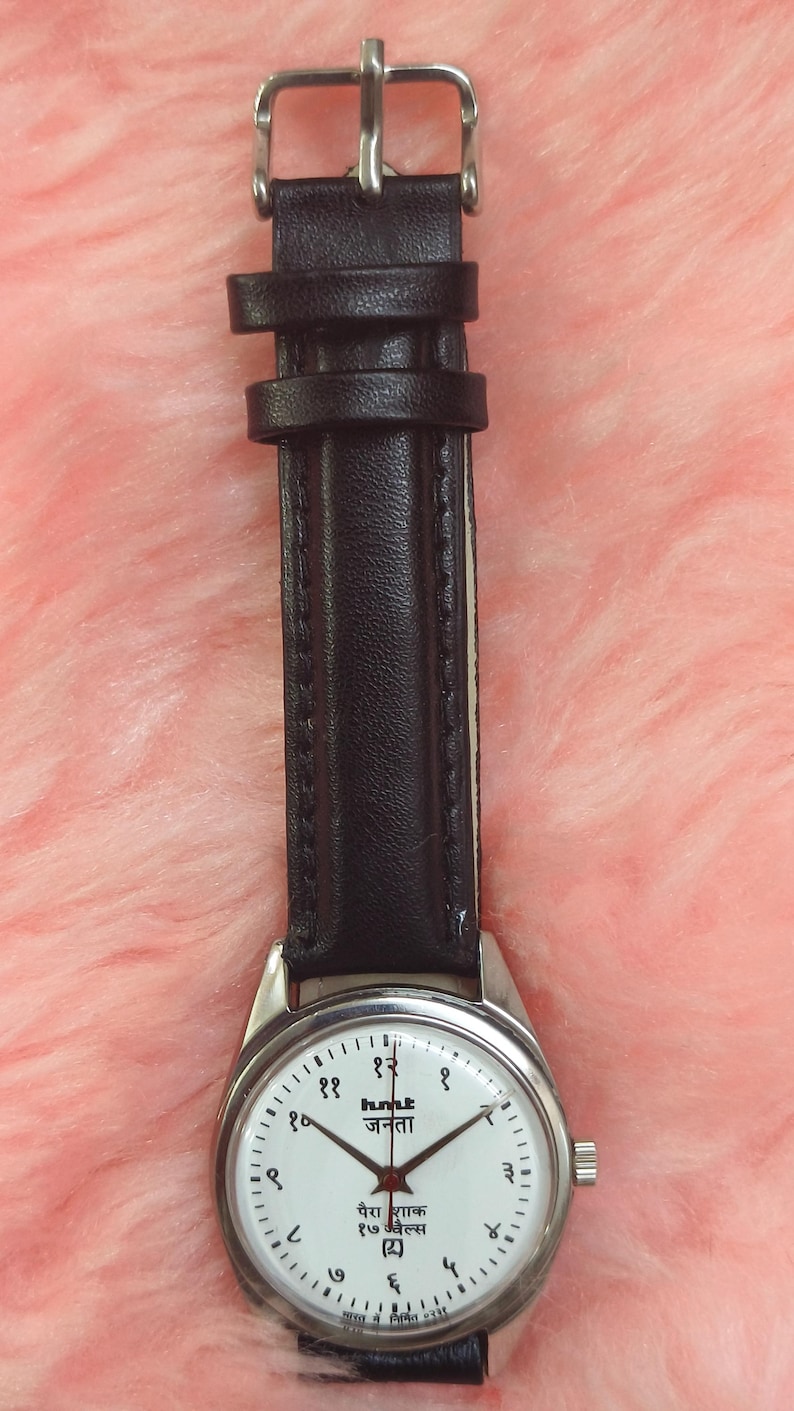 May include: A wristwatch with a silver-colored case and a black leather strap. The watch face is white with black numerals and text. The watch has a buckle closure.