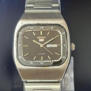 May include: A silver-tone Seiko 5 automatic wristwatch with a square-shaped case and a matching metal link bracelet. The dark brown dial features silver hour markers, a day-date window at the three o'clock position, and a 24-hour scale around the edge.