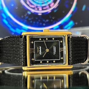 May include: A rectangular gold-tone wristwatch with a black dial featuring gold-tone hour markers, gold hands, and the text HMT Ashwini, Para Shock, 17 Jewels. The watch has a black woven fabric strap with silver-tone metal hardware loops.