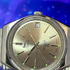 Op de afbeelding: Zilverkleurig Tarzeeg polshorloge met een lichtgekleurde wijzerplaat, zwarte en groene accenten. Het horloge heeft een datumvenster en de tekst "PARA SHOCK 17 JEWELS". De horlogeband is zilverkleurig en het horloge staat tegen een blauwe achtergrond.