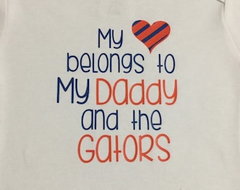 May include: White baby onesie with the text "My belongs to My Daddy and the GATORS" in blue and orange. A heart with blue and orange stripes is above the text. The onesie has short sleeves.