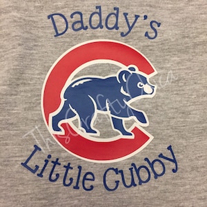 May include: A gray onesie with the text "Daddy's Little Cubby" in blue lettering. A red and blue Chicago Cubs logo with a bear is in the center of the onesie.