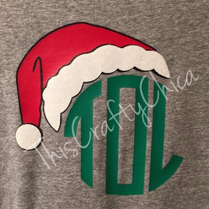 Puede incluir: Una camiseta gris con un monograma verde y un gorro de Papá Noel rojo y blanco.