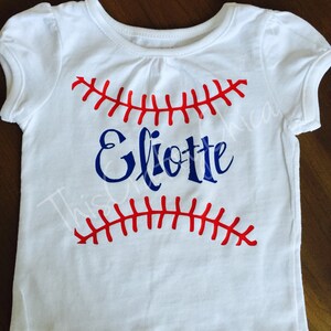 Op de afbeelding: Wit T-shirt met een rood en blauw baseball ontwerp en de naam "Eliotte" in blauwe letters.