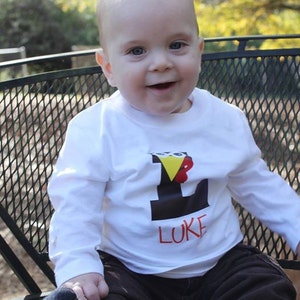 Könnte beinhalten: Ein kleiner Junge trägt ein weißes Langarmshirt mit einem schwarzen und roten Buchstaben L und dem Namen "LUKE" darauf. Er sitzt auf einem schwarzen Metallstuhl mit einem Netzdesign.