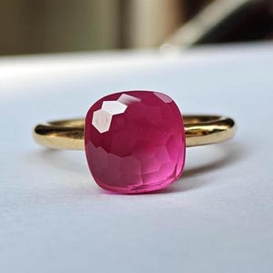 May include: A gold ring featuring a large, faceted, square-cut, magenta gemstone. The ring's band is a simple, polished gold. The gemstone has a high degree of clarity and reflects light, creating a sparkling effect. The ring is set against a neutral background.