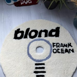 May include: A round, off-white rug with the word "blond" in black block letters and "FRANK OCEAN" in black text. A light blue circle surrounds a central hole. Black lines form a design below the circle. A smaller rug is visible in the background.