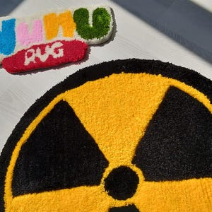 May include: A round, yellow and black rug featuring a radiation symbol. Above it, a smaller rug spells out "NIKI RUG" in various colors. The rugs are placed on a light-colored surface.