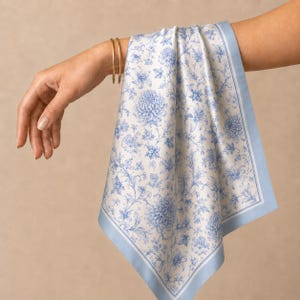 Op de afbeelding: Een vierkante zijden sjaal met een witte achtergrond en een herhalend blauw bloemenpatroon van chrysanten en lelies. De sjaal is afgewerkt met een effen lichtblauwe rand en een dunne witte binnenrand voor een klassieke en elegante uitstraling.