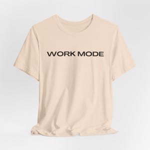 Könnte beinhalten: Hellbeige T-Shirt mit dem schwarzen Schriftzug "WORK MODE" auf der Brust. Das Shirt hat kurze Ärmel und einen Rundhalsausschnitt. Der Stoff wirkt weich und leicht zerknittert.