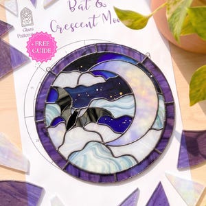 Könnte beinhalten: Ein kreisförmiger Buntglas-Sonnenfänger mit einer Fledermaus-Silhouette vor einem Halbmond und nächtlichem Himmel. Das Stück besteht aus violetten, blauen und weißen Glassegmenten mit dunklen Bleilinien. Es liegt auf einer Papiervorlage mit Muster.