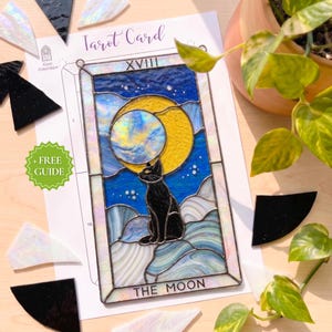 Può includere: Un pannello rettangolare in vetro colorato raffigurante un gatto nero seduto sotto una mezzaluna gialla e un grande globo iridescente su un cielo blu. Il pezzo presenta il numero romano XVIII e il testo THE MOON in una cornice di metallo scuro.