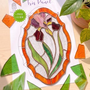 Peut inclure: Un panneau en vitrail ovale représentant une fleur d'iris violette et rose avec des feuilles vertes, entouré d'une bordure orange. Le panneau repose sur un patron en papier blanc intitulé Iris Panel avec une icône verte + FREE GUIDE.