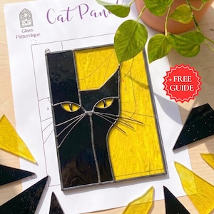 May include: A stained glass cat panel with a black and yellow design. The cat's face is black, with yellow eyes and whiskers, set against a yellow background. The panel is on a white paper with the words "Cat Panel" and "Glass Patternique".