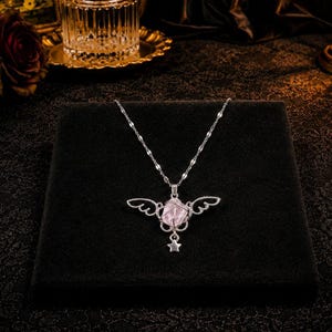 May include: A silver-colored necklace with a pendant featuring a pink gemstone, delicate wing-shaped accents, and a small star charm. The necklace is displayed on a black surface, with a blurred background.
