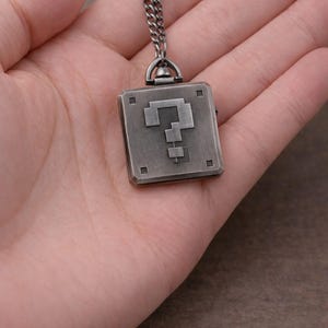 May include: A silver-coloured pendant necklace featuring a pixelated question mark design. The pendant is square-shaped with a chain attached. The necklace is held in a hand, showcasing the details of the pendant's design.