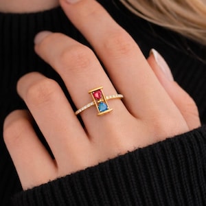 Puede incluir: Un anillo dorado con un diseño rectangular que presenta una gema roja y azul. La banda del anillo está adornada con pequeñas gemas transparentes. El anillo es una joya.