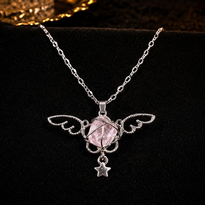 May include: A silver-toned necklace featuring a pink crystal pendant encased in a wireframe design with wing accents. A small star charm dangles below the crystal. The necklace is set against a dark background.
