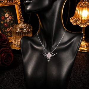 May include: A silver necklace with a pendant featuring a pink rose design with wing accents and a small star charm. The necklace is displayed on a black mannequin, with a blurred background of decorative items.