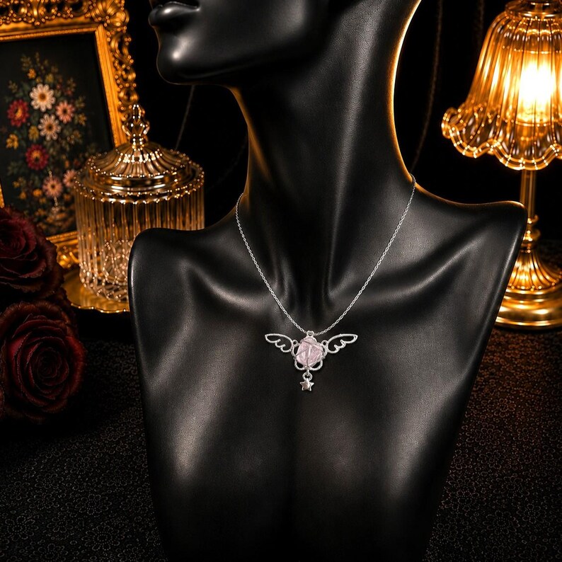 May include: A silver necklace with a pendant featuring a pink rose design with wing accents and a small star charm. The necklace is displayed on a black mannequin, with a blurred background of decorative items.