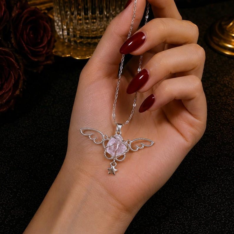 May include: A silver necklace with a pendant featuring a pink crystal, wing-shaped filigree, and a small star. The necklace is held in a hand with red nail polish, against a dark background.