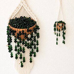 Peut inclure: Deux décorations végétales suspendues composées de pots en terre cuite remplis de perles de cristal facettées vert foncé qui retombent. Le plus grand pot est suspendu par un cordon en macramé crème à un anneau en bois, le plus petit par une chaînette.