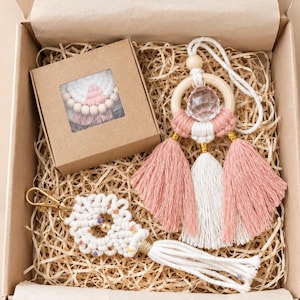 Peut inclure: Une boîte cadeau en carton contenant une petite boîte carrée avec une fenêtre, un porte-clés en macramé avec cordon blanc et détails dorés, et une suspension avec anneau en bois, cristal facetté et pompons en coton rose et blanc.