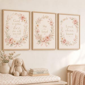 Op de afbeelding: Drie ingelijste kunstprints met bloemenkransontwerpen en inspirerende citaten. De prints hebben zachte roze en crèmekleuren, met tekst die luidt: "God is in haar, zij zal niet vallen", "Zij is geliefd" en "Geliefd, gekozen, bekend".
