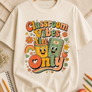 Könnte beinhalten: Cremefarbenes T-Shirt mit einem farbenfrohen Aufdruck "Classroom Vibes Only". Das Design umfasst einen Stiftebecher, ein Notizbuch, einen Regenbogen, Blumen und Sterne. Das T-Shirt hängt an einem Holzbügel.