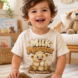 Op de afbeelding: Een beige t-shirt met de tekst "Milk Monster" en een cartoonmonster met een melkfles. De tekst "Highly Volatile Until Fed" staat eronder. Het kind draagt een bruine broek.