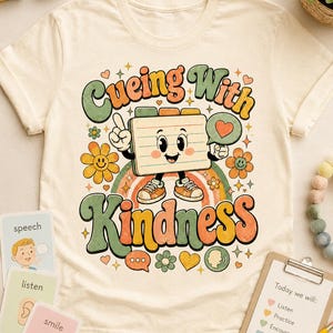 Op de afbeelding: Crèmekleurig T-shirt met een kleurrijke afbeelding met de tekst "Cueing With Kindness". Het ontwerp toont een lachend notitieblokkarakter, bloemen en een regenboog. Extra kaarten en een klembord met tekst zijn zichtbaar.