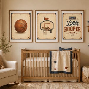 May include: Three framed basketball-themed prints. The first shows a detailed basketball, the second a hoop with a flag, and the third reads "Little Hooper." The prints have a vintage aesthetic, with a cream background and brown frames. The room also features a crib and a rocking chair.