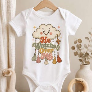 May include: White baby bodysuit with a retro design featuring a smiling cloud, stars, and the text "He Watches Over Me." The design includes colorful tassels and a cross pendant, perfect for a baby's wardrobe.