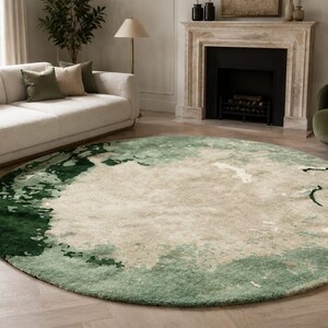 May include: A large round area rug featuring an abstract pattern with a central cream-colored field surrounded by blended gradients of sage green and deep forest green, creating a textured, layered appearance across the circular surface.