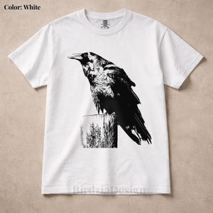 Può includere: T-shirt bianca con un'illustrazione in bianco e nero di un corvo appollaiato su un palo di legno. Il corvo è raffigurato in uno stile artistico dettagliato. Il testo "Birdzia Design" è nella parte inferiore della maglietta.