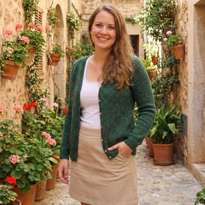 May include: A woman wearing a green cardigan with a white top and a tan skirt. The cardigan has small flecks of color throughout. The woman is smiling and standing in a stone alleyway with potted plants.
