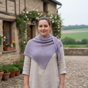 May include: A woman wearing a lavender knitted shawl. The shawl is triangular, with a textured pattern and a decorative openwork section near the neck. She is standing in front of a stone building with climbing roses and potted plants.